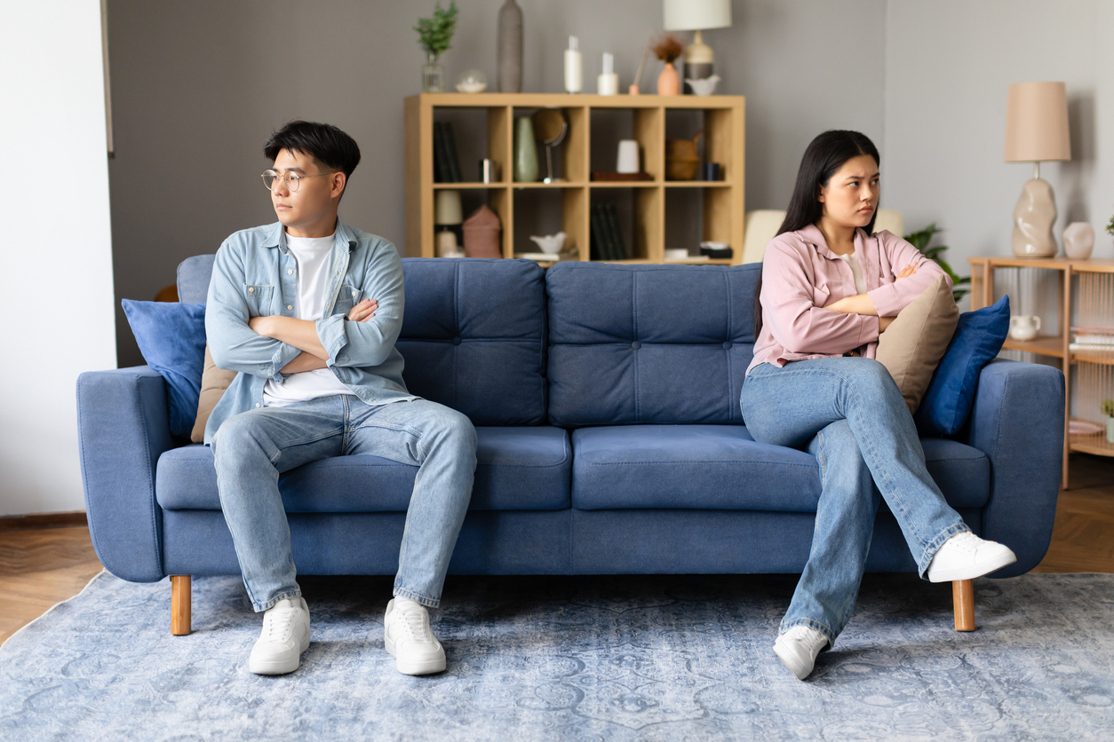 Korean couple sitting upset and offended, distancing themselves after argument in the living room, looking at sides and thinking about divorce. Relational challenges and unresolved conflicts